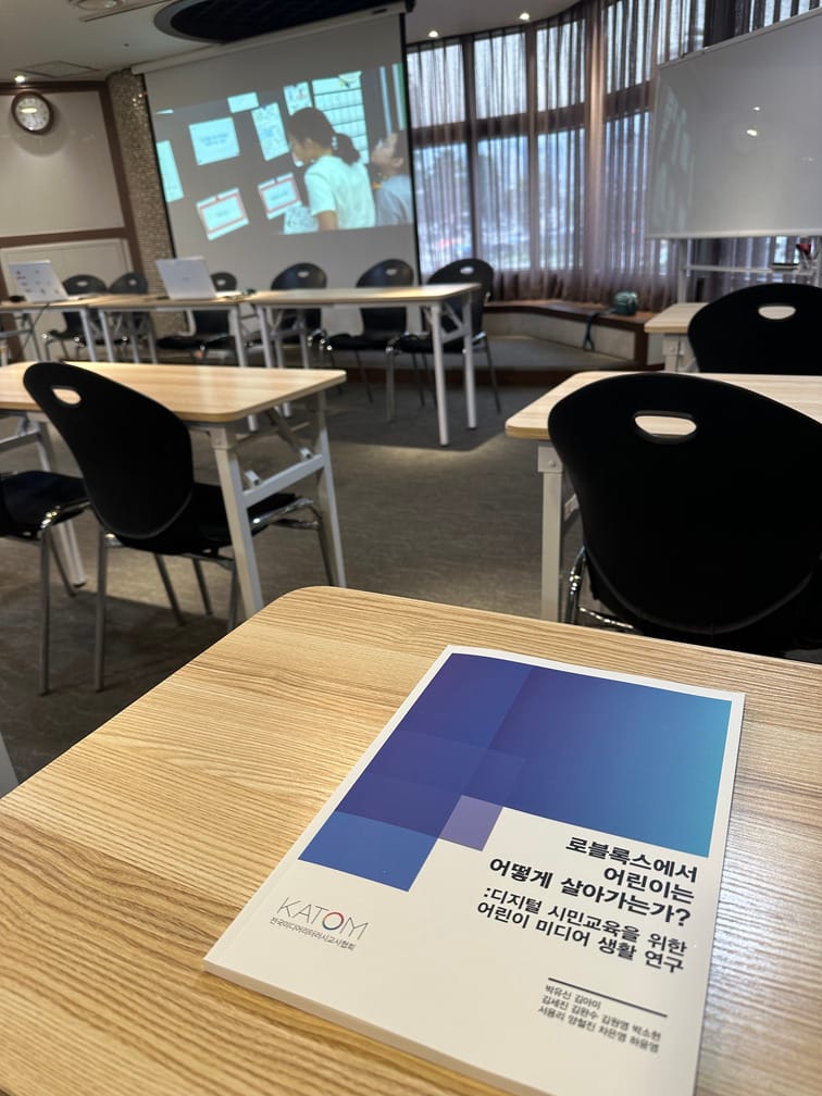 강의실의 책상에 『로블록스에서 어린이는 어떻게 살아가는가?』라는 제목의 연구집이 놓여있고, 단상쪽 모니터에는 아이가 칠판에 붙은 종이를 향해 선 영상이 틀어져 있다.