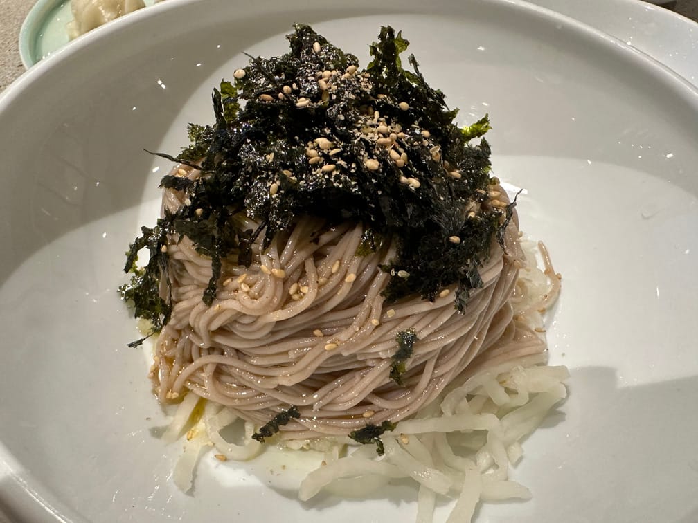 흰 면기 가운데에 흰 무채, 들기름과 깨가 비벼진 메밀면, 김가루, 다시 깨가루가 순서대로 쌓여 있다.