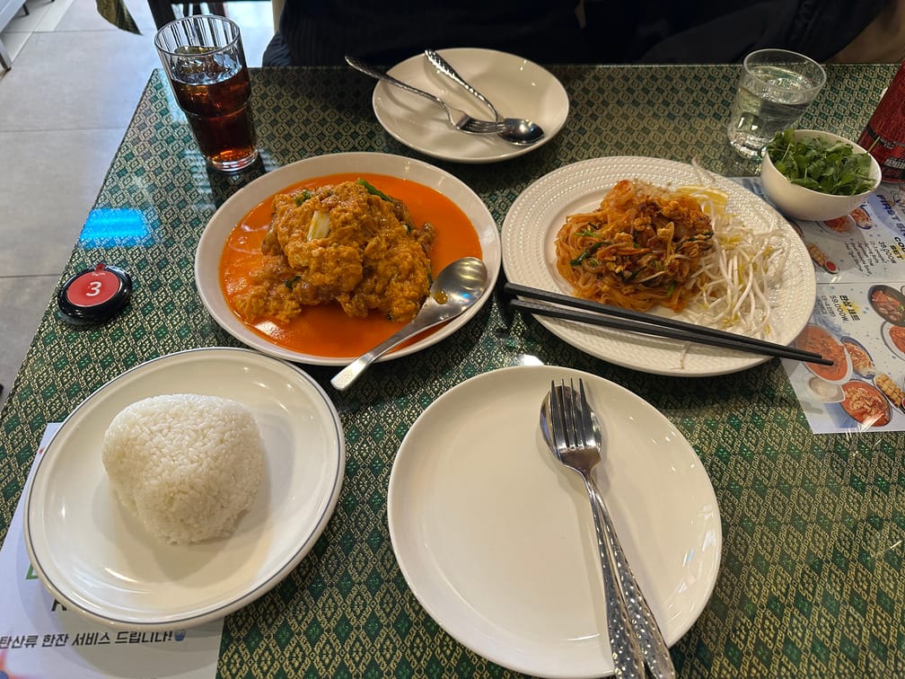 테이블 위에 붉은 국물과 계란으로 덮인 요리, 붉은 볶음국수와 숙주나물, 그릇에 담긴 고수, 접시 위의 하트모양 밥이 차려져 있다.