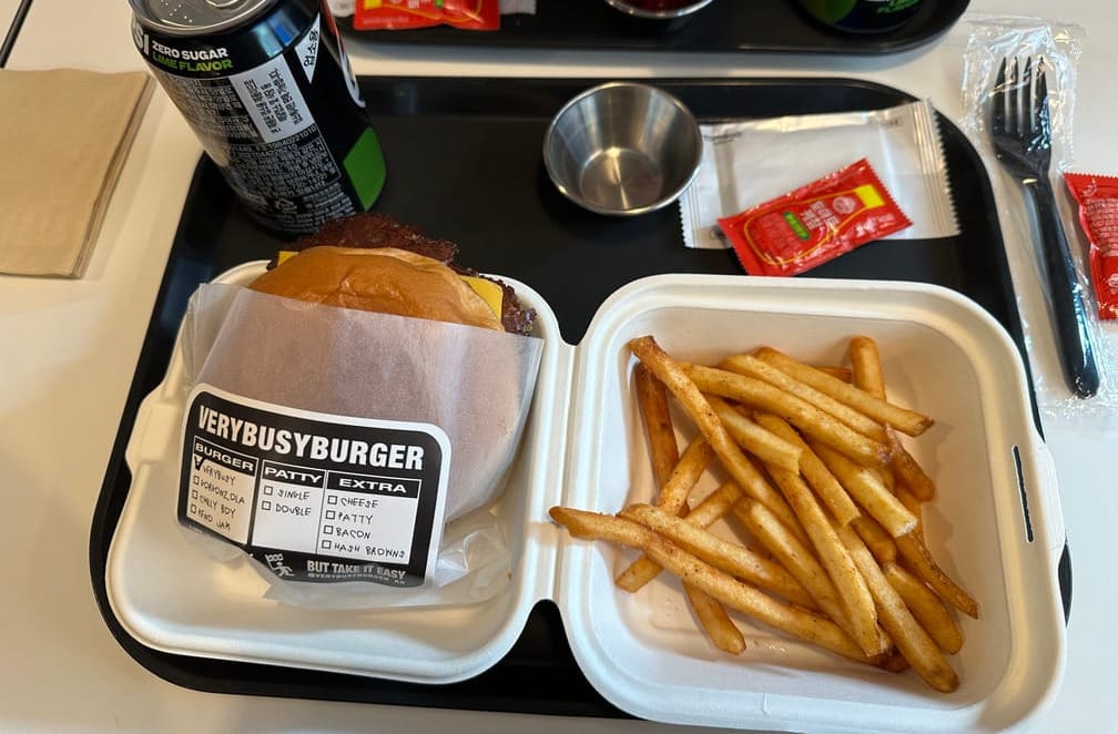 검은 트레이에 종이 버거 상자가 있고 그 안에 VERYBUSYBURGER라고 적힌 스티커가 붙은 봉투 안에 담긴 버거와 감자튀김이 보이는 사진. 트레이 위에는 콜라, 케찹, 케찹 용기 등도 보인다.