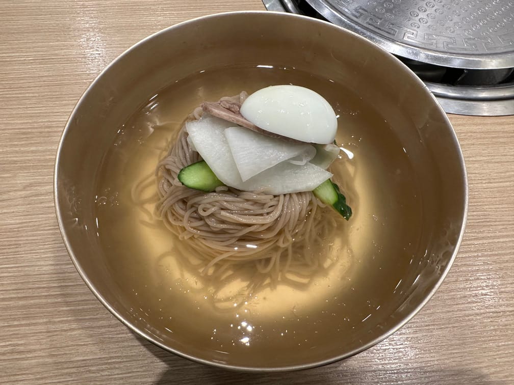 면기에 맑은 국물, 메밀면, 오이, 무절임, 고기, 달걀 반쪽이 담겨 있는 냉면을 내려다본 사진.