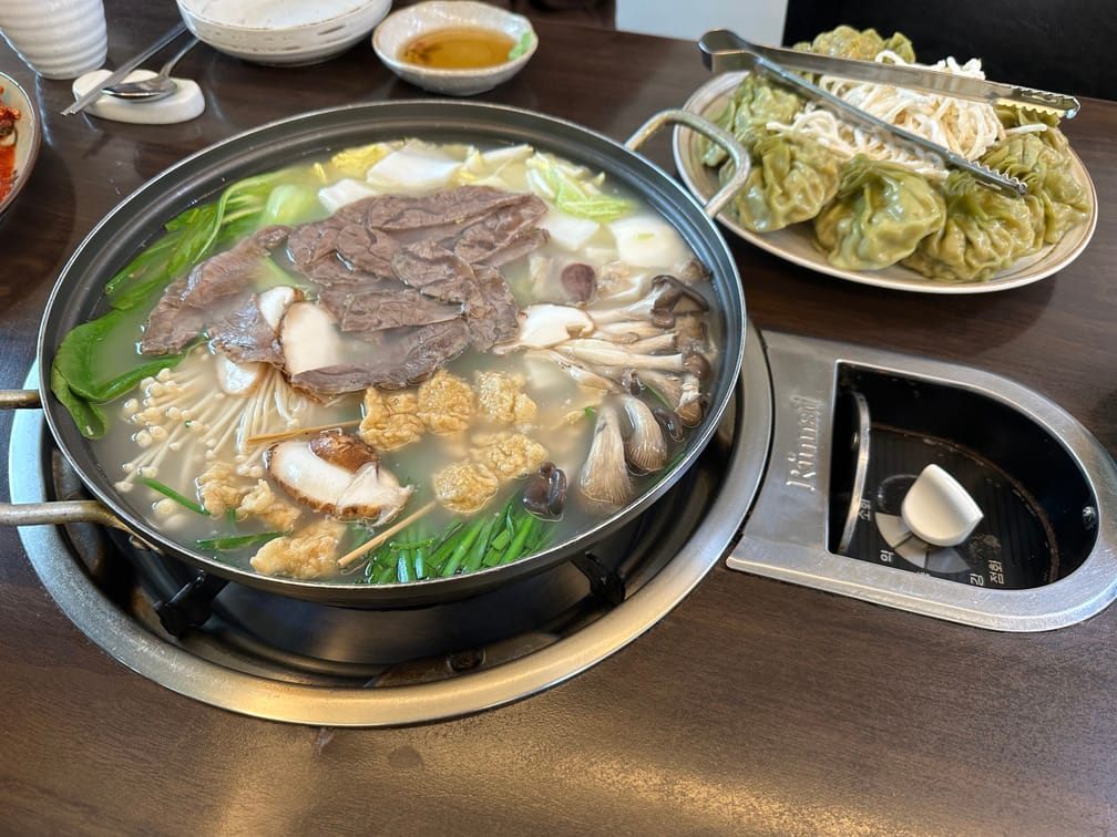 가스레인지 식탁에 전골 냄비가 올라가 있고 뿌연 국물 전골에 배추, 청경채, 팽이버섯, 느타리버섯, 부추, 얇은 고기 등이 들어가 있고 접시에 만두와 칼국수가 차려져 있는 모습.