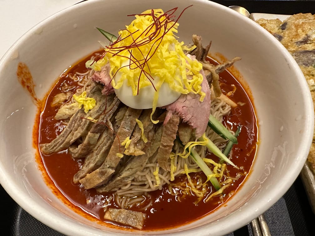 흰 그릇에 담긴 붉은 양념과 면, 오이, 육전, 수육, 계란 반쪽 계란 지단, 실고추.