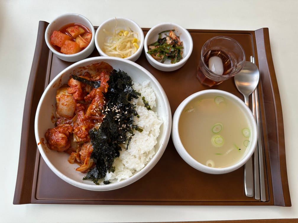 식탁에 나무쟁반이 올라와 있고, 김가루와 붉은 양념의 닭고기가 올라간 밥과 뿌연 국물, 깍두기, 콩나물무침, 부추김치와 매실차가 차려져 있다.