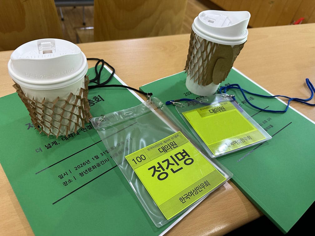 좌우로 긴 책상에 얇은 녹색 책자, 연두색 명찰, 종이컵이 각각 두 개씩 놓여 있는 사진.