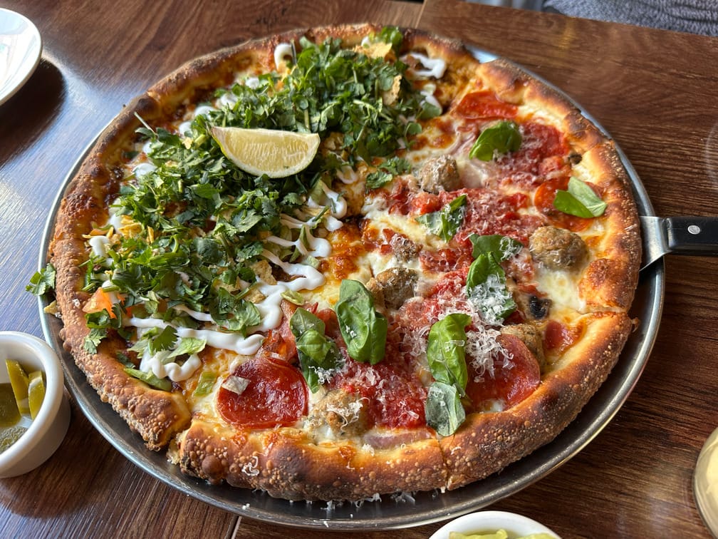 나무 테이블에 올라온 갓 구워진 피자. 페퍼로니가 올라간 피자와 고수로 뒤덮여있고 라임이 올라와있는 피자의 반반 조합.