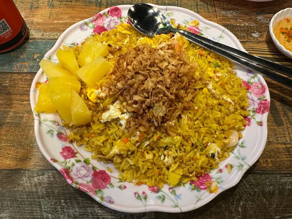 나무 테이블 위 꽃무니 그릇에 노란 볶음밥과 파인애플, 찢어진 달걀 후라이, 후레이크가 올라가 있는 사진